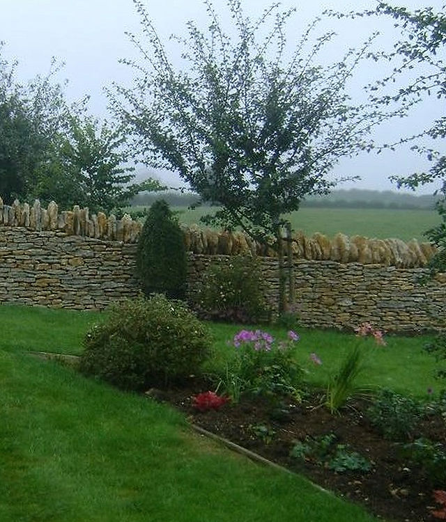 A dry stone wall beside a lawn and planted border, with fields beyond