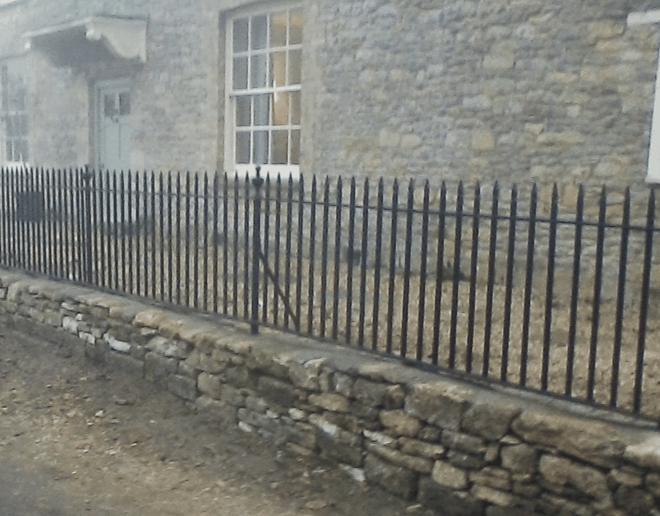 Black iron spear-top railings above a low stone wall in front of a stone house