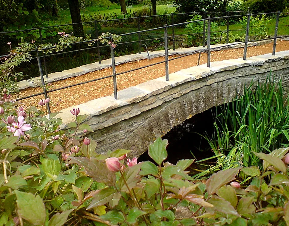 A curved stone bridge over a stream in a planted garden
